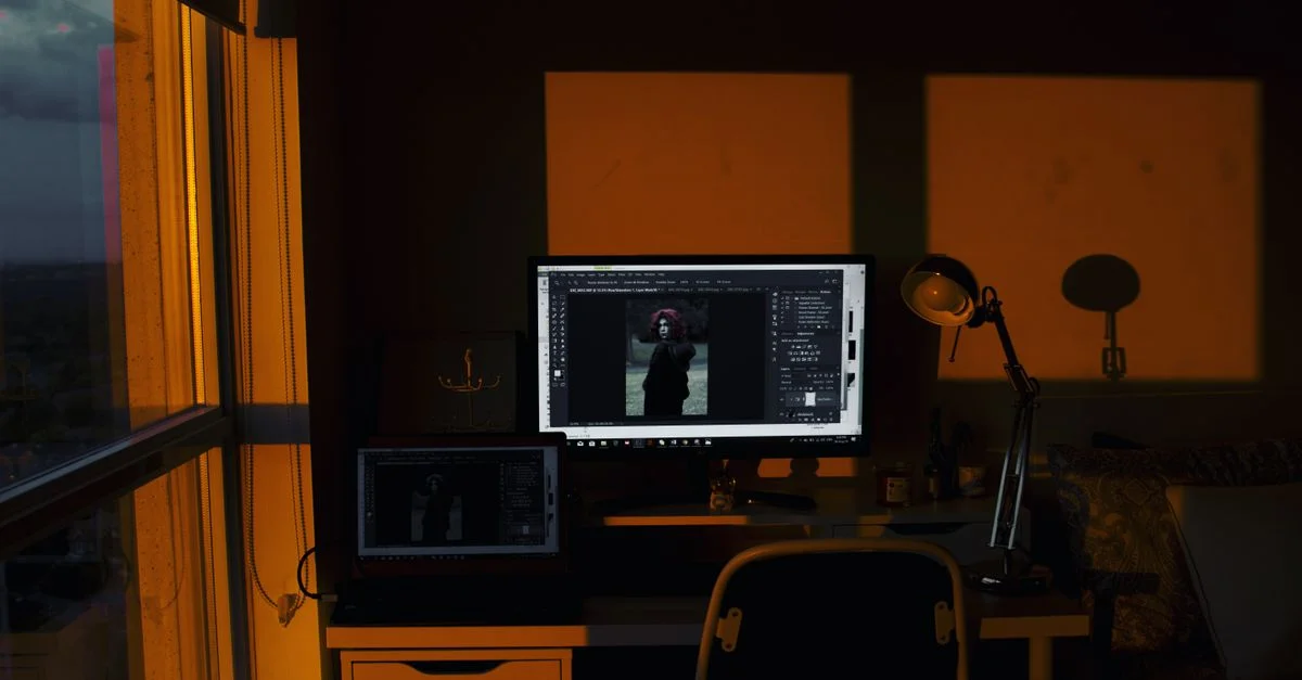 Un bureau à domicile faiblement éclairé avec un ordinateur de bureau et un ordinateur portable illuminés par la lumière orange du coucher de soleil.