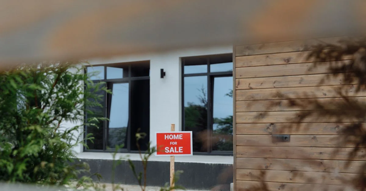 Une maison contemporaine avec un panneau 'À Vendre' dans le jardin avant, encadrée par de la verdure.