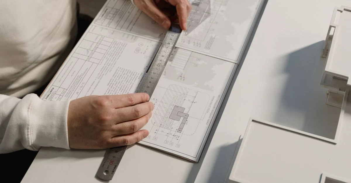Mains d'une personne dessinant des plans architecturaux avec une règle sur papier, à l'intérieur.