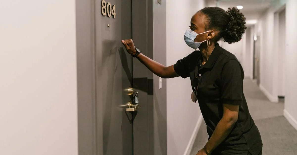 Une femme de livraison portant un masque frappe à une porte dans un couloir intérieur, promouvant la sécurité et la logistique.